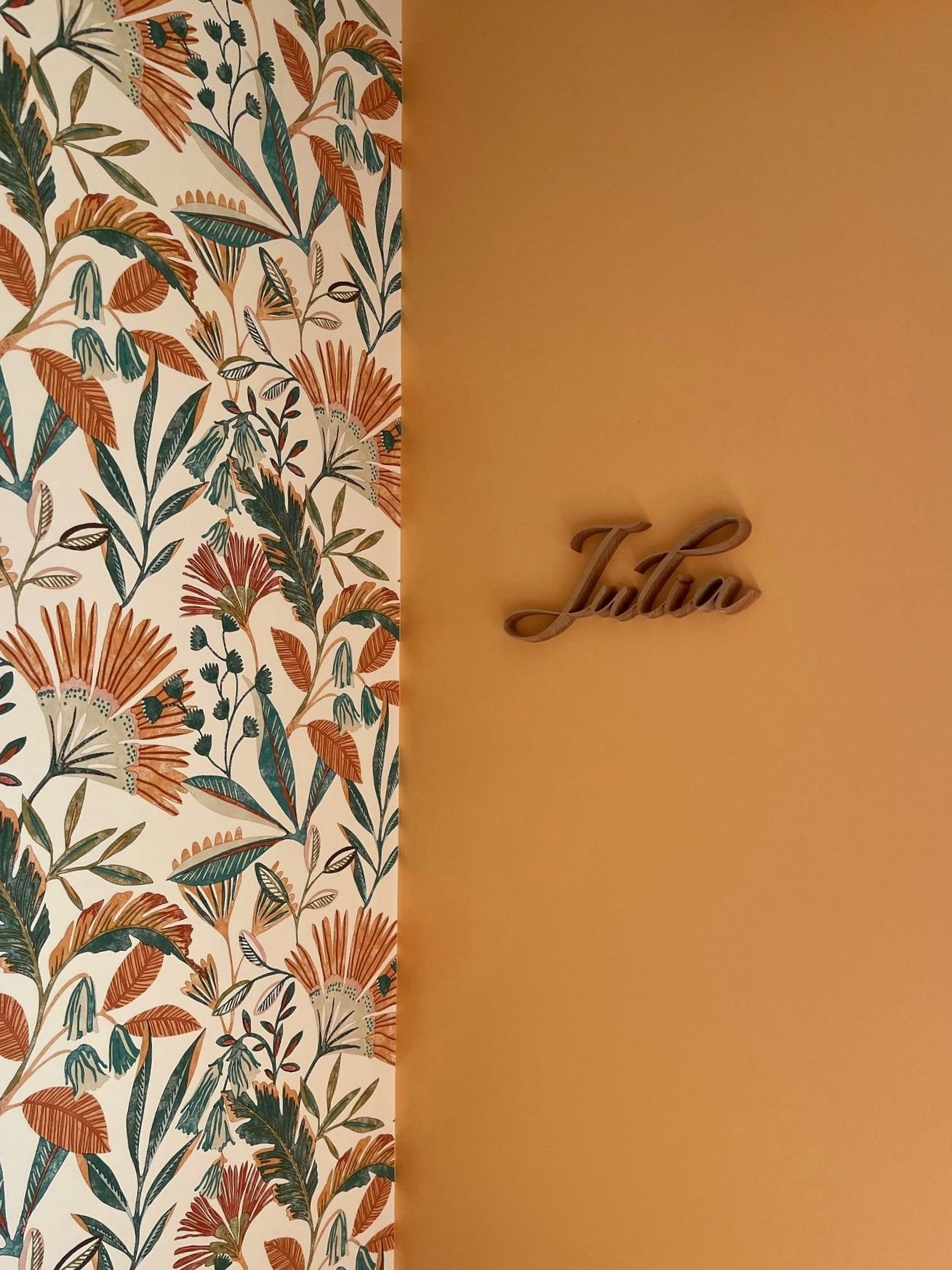 Prénom Julia en bois fixé sur un mur ocre à côté d'un papier peint fleuri